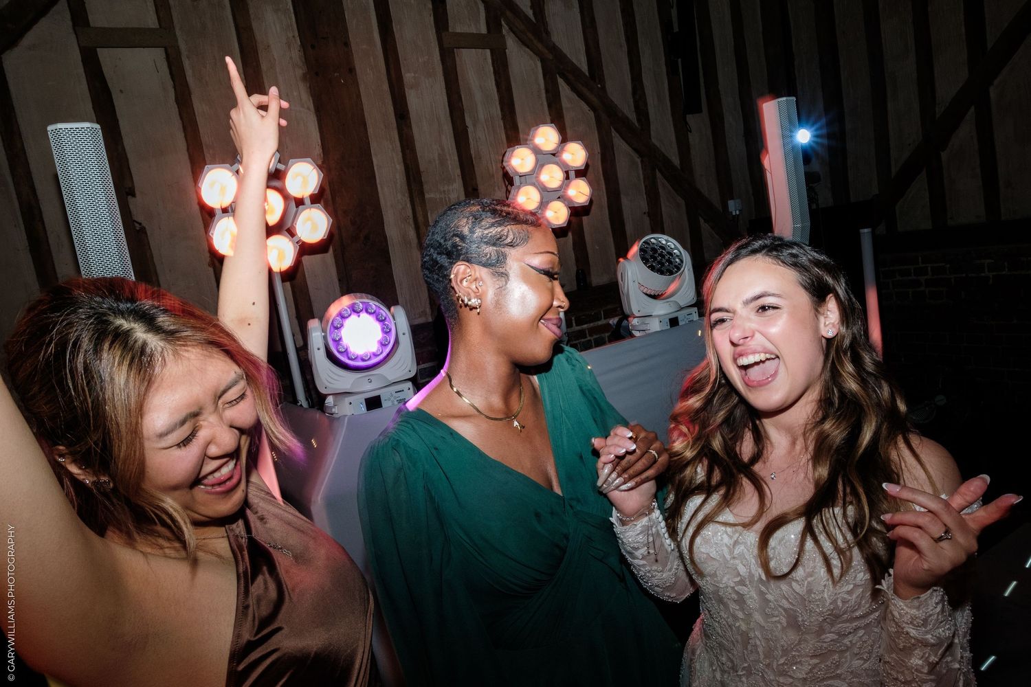 Three women dancing joyfully at a wedding reception with colorful stage lights in the background.