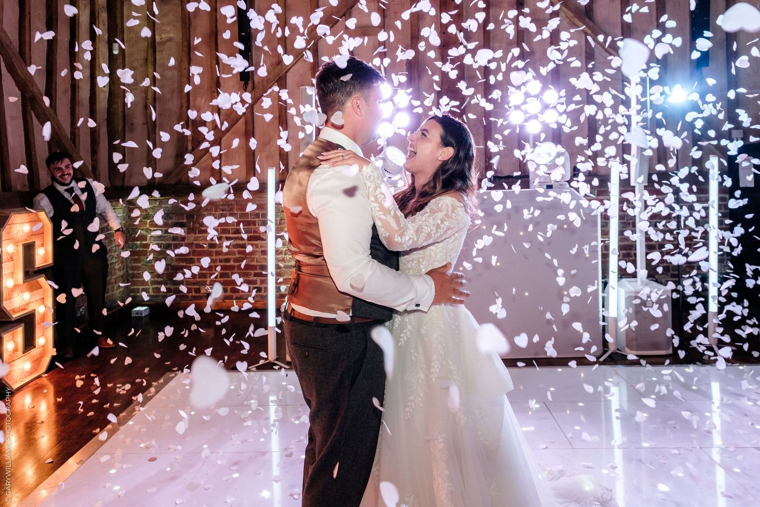 Bride and groom share first dance surrounded by falling white confetti in a warmly lit rustic wedding venue.