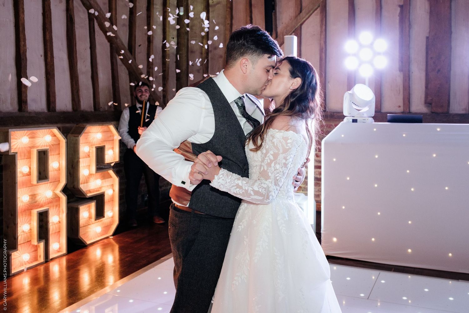 Bride and groom sharing first dance kiss at wedding reception with illuminated RS marquee letters in rustic barn venue.