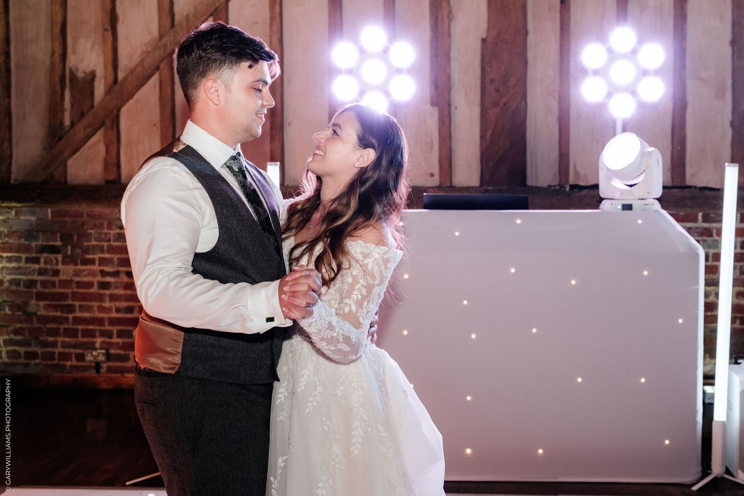A bride and groom share their first dance at a wedding reception in a rustic barn venue with bright stage lighting.