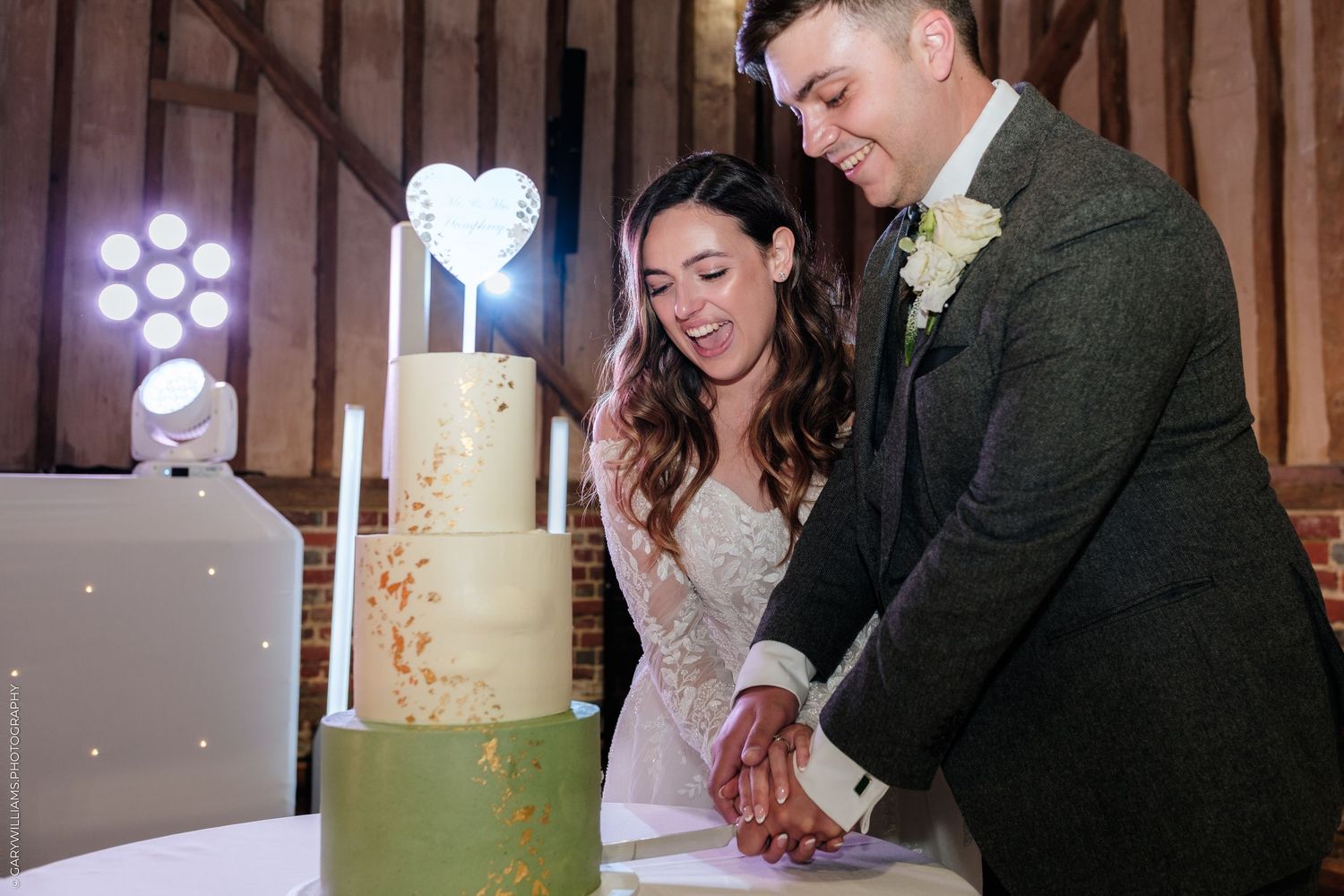 Bride and groom cutting a three-tier green and gold wedding cake in a rustic barn venue.