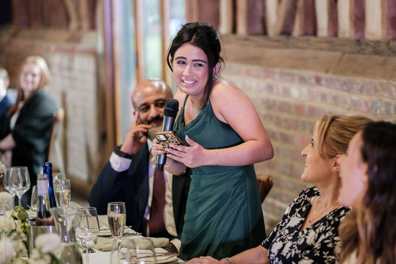Woman in green dress giving a speech at an elegant wedding reception dinner in a rustic barn venue.