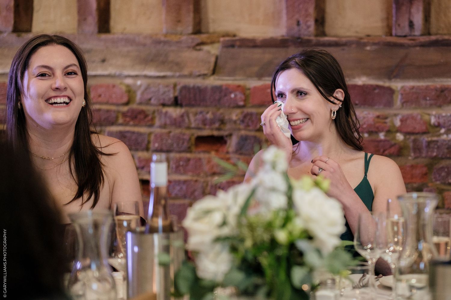 Two women laughing and crying happy tears at a wedding reception dinner, seated at a decorated table with floral centerpieces.