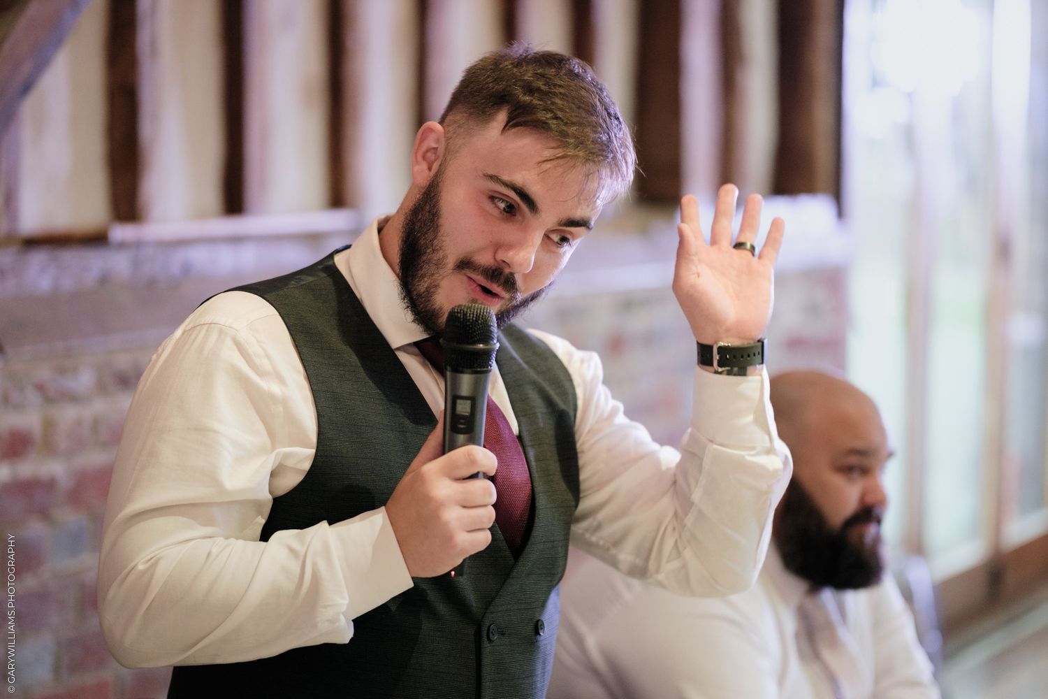 A bearded man in a vest holds a microphone and raises his hand while speaking at a wedding reception.