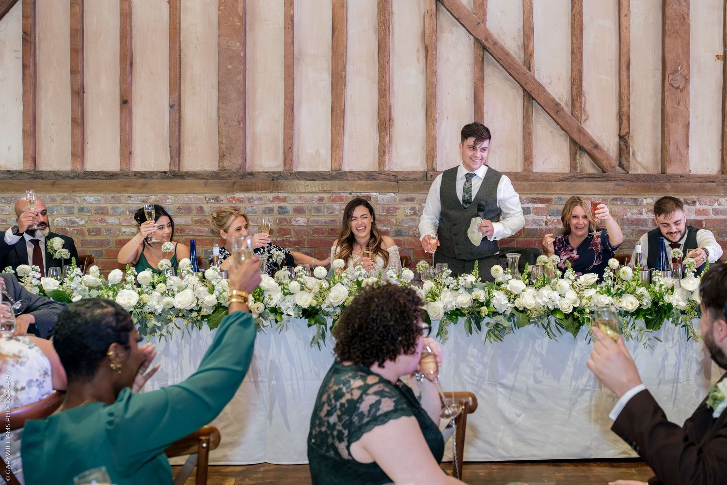 Wedding reception toast in rustic barn venue with floral arrangements and guests raising glasses to celebrate.
