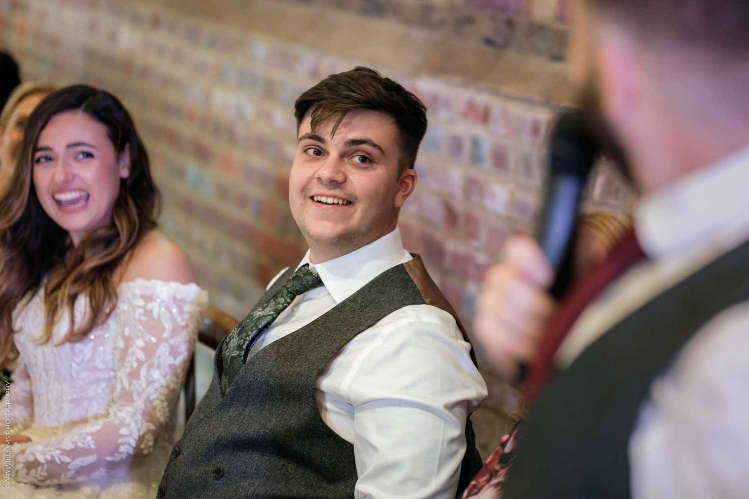 Smiling groom in a grey waistcoat listening to a wedding speech at a rustic brick-walled reception venue.