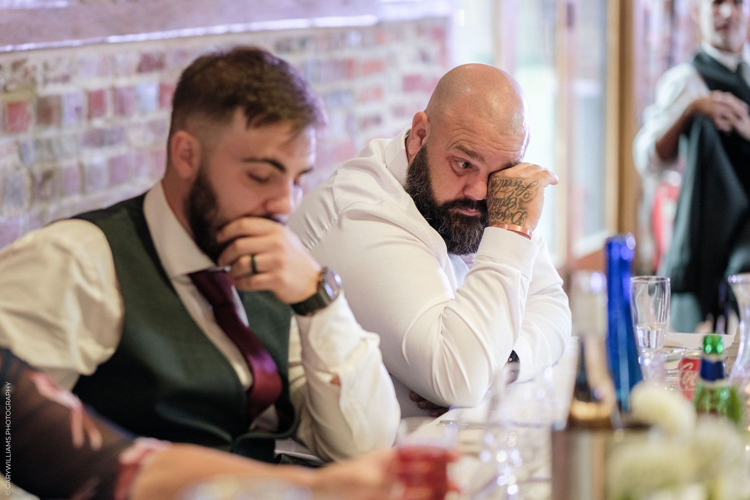 Two bearded men in formal attire sitting at a table, appearing deep in thought at a wedding reception.