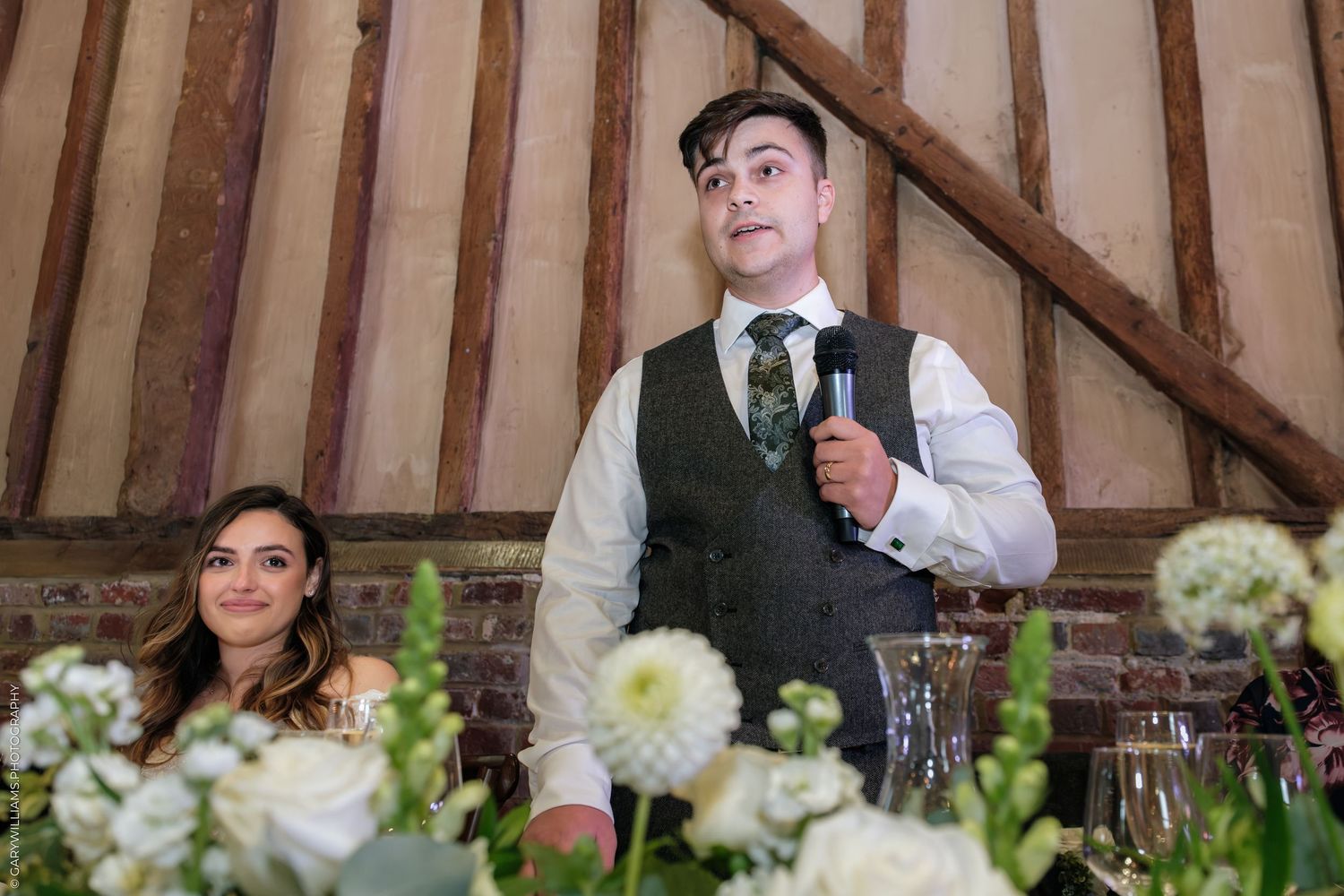 Groom delivering wedding speech at rustic barn venue with bride seated beside floral arrangements.