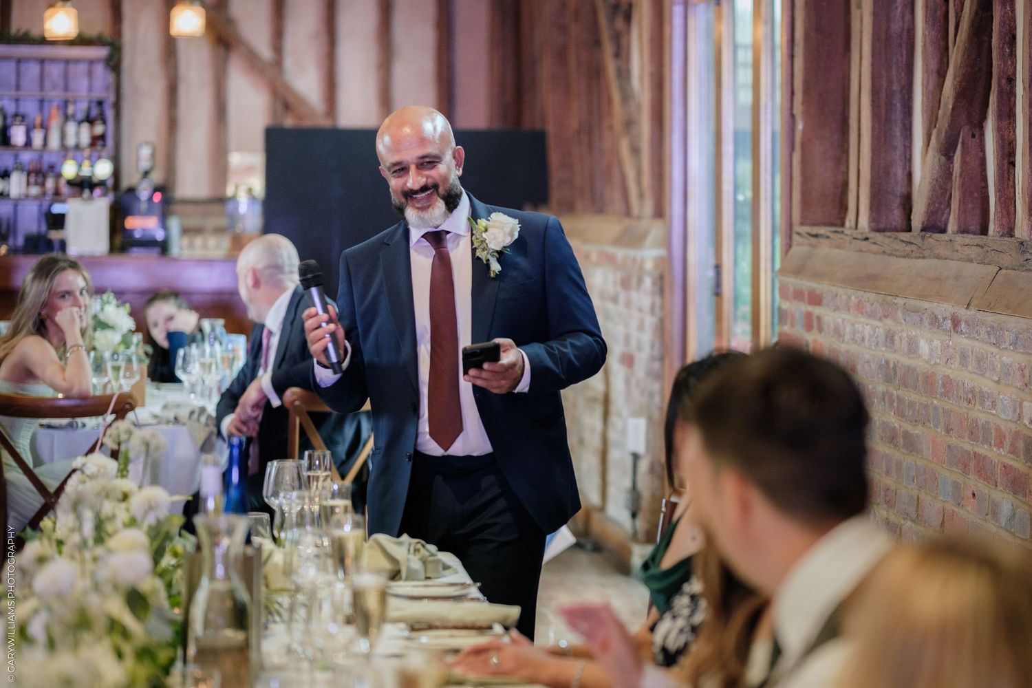Man in navy suit giving a speech at an elegant wedding reception dinner in a rustic barn venue.