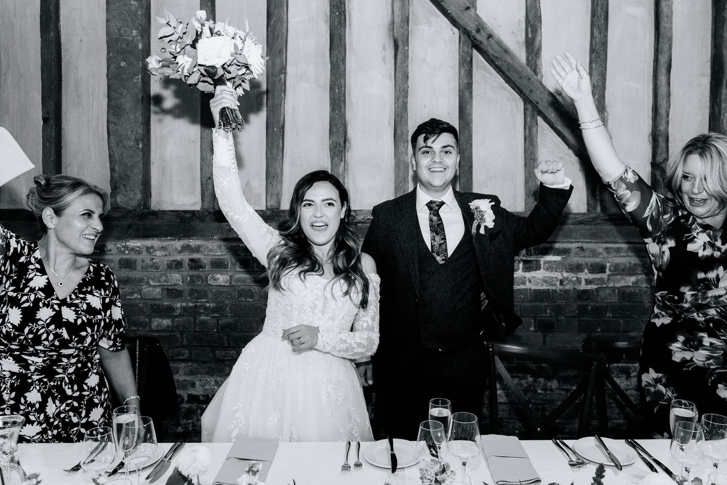 Black and white photo of a joyful bride and groom raising their arms at wedding reception table.