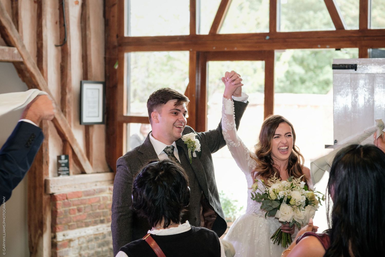 Joyful newlyweds raise hands in celebration at rustic barn wedding venue, bride holding white bouquet surrounded by guests.