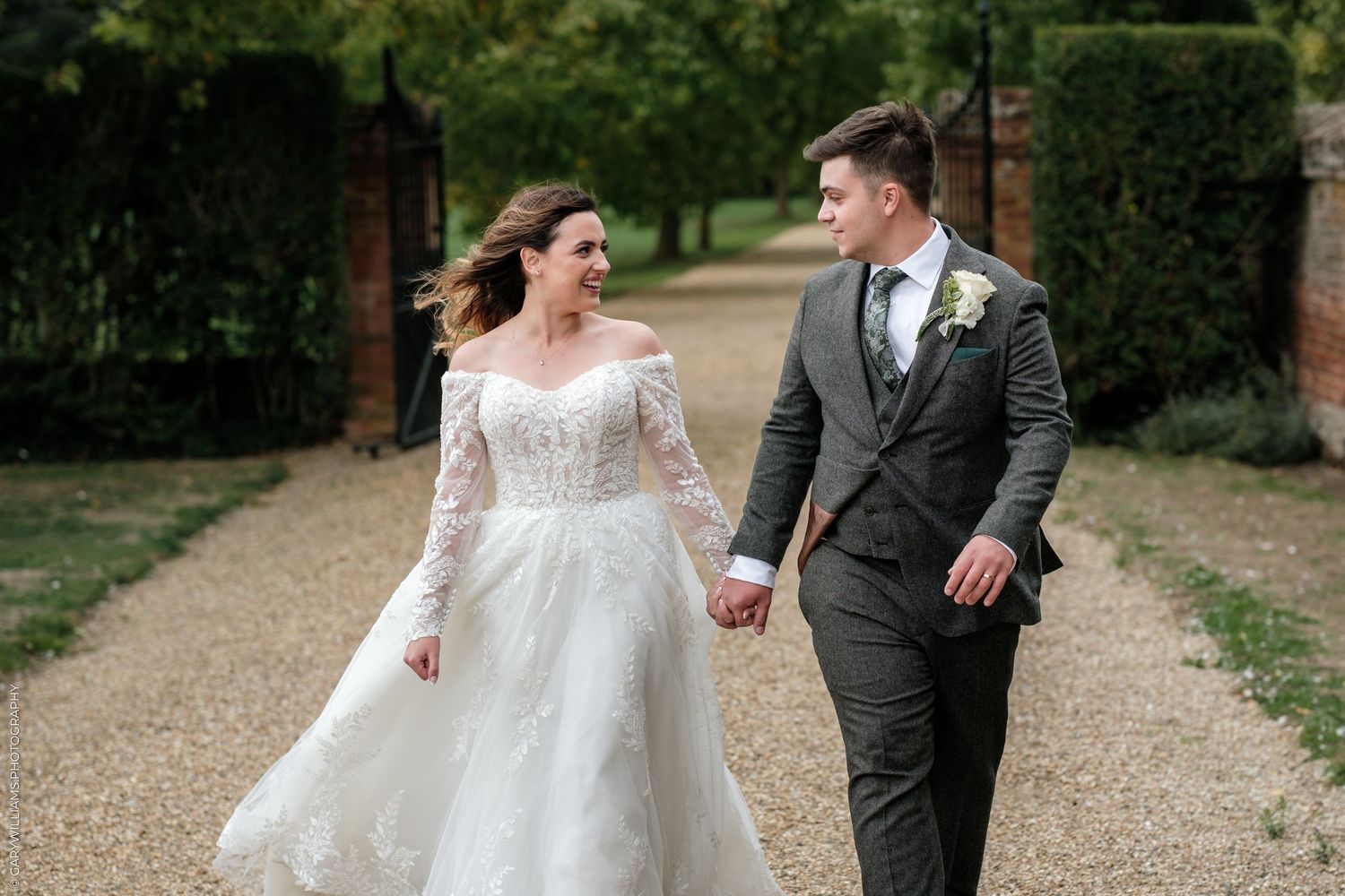 A bride in a lace off-shoulder gown and groom in grey suit hold hands walking along a garden path.