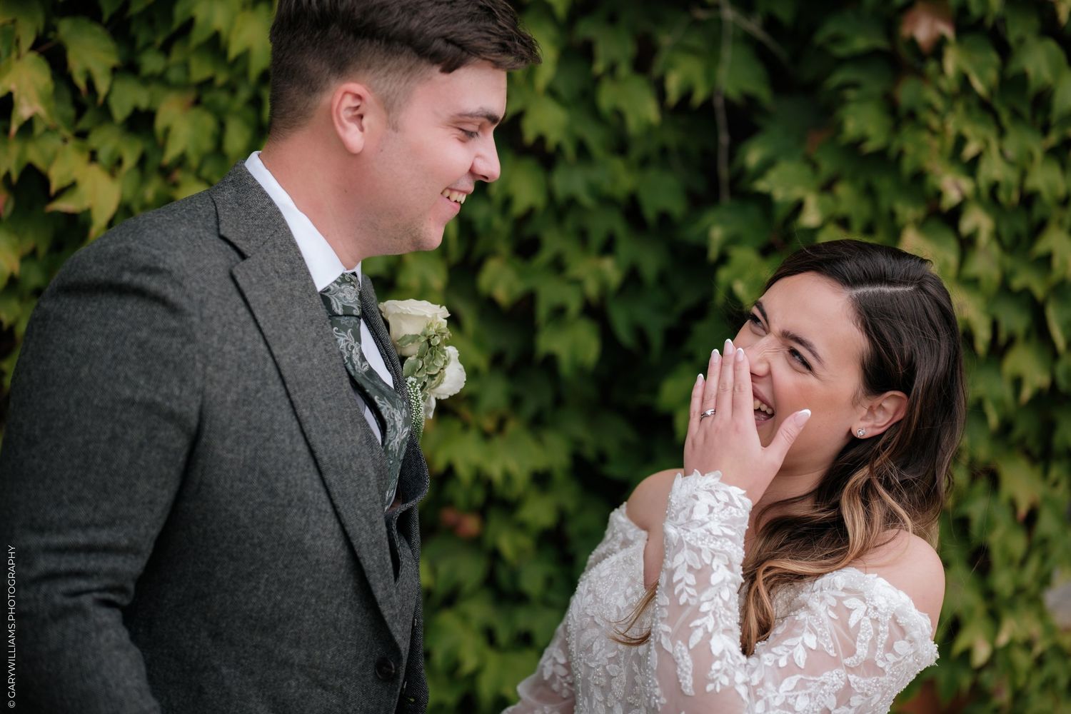 Bride laughing joyfully with groom in grey suit, standing before lush green ivy wall at wedding.