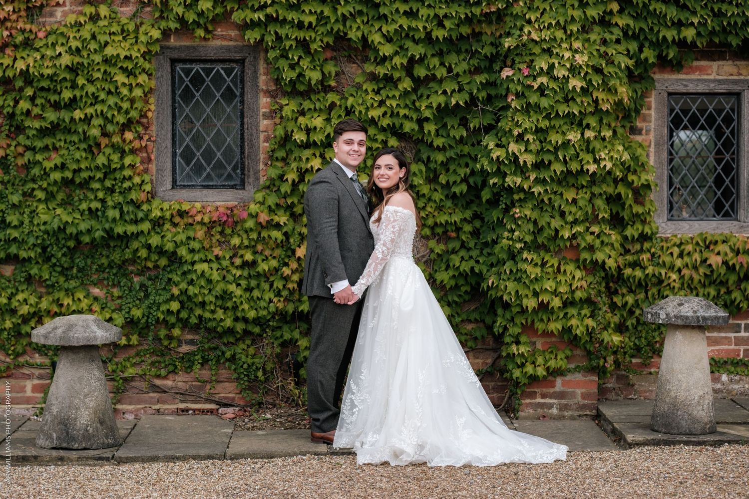 Bride and groom holding hands posing before ivy-covered brick wall at elegant outdoor wedding venue.