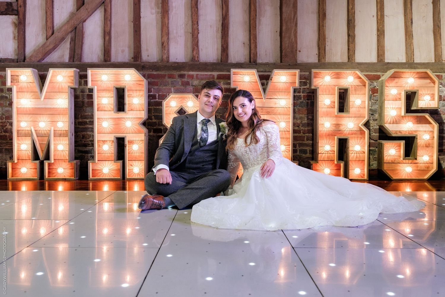 Newlywed couple sitting on dance floor in front of illuminated Mr & Mrs marquee letters at wedding reception.