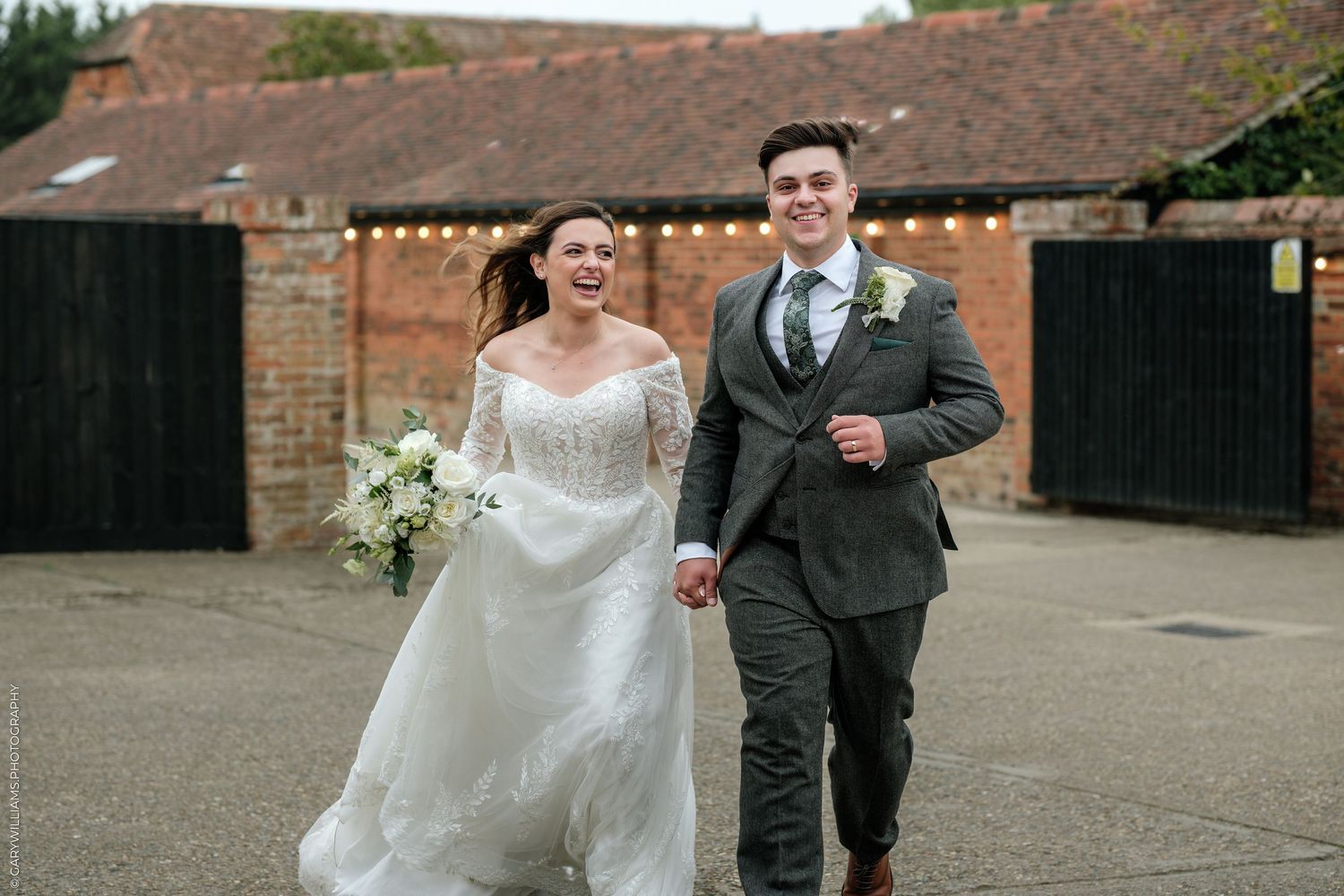 Happy bride and groom walking hand in hand outside a rustic barn wedding venue decorated with fairy lights.