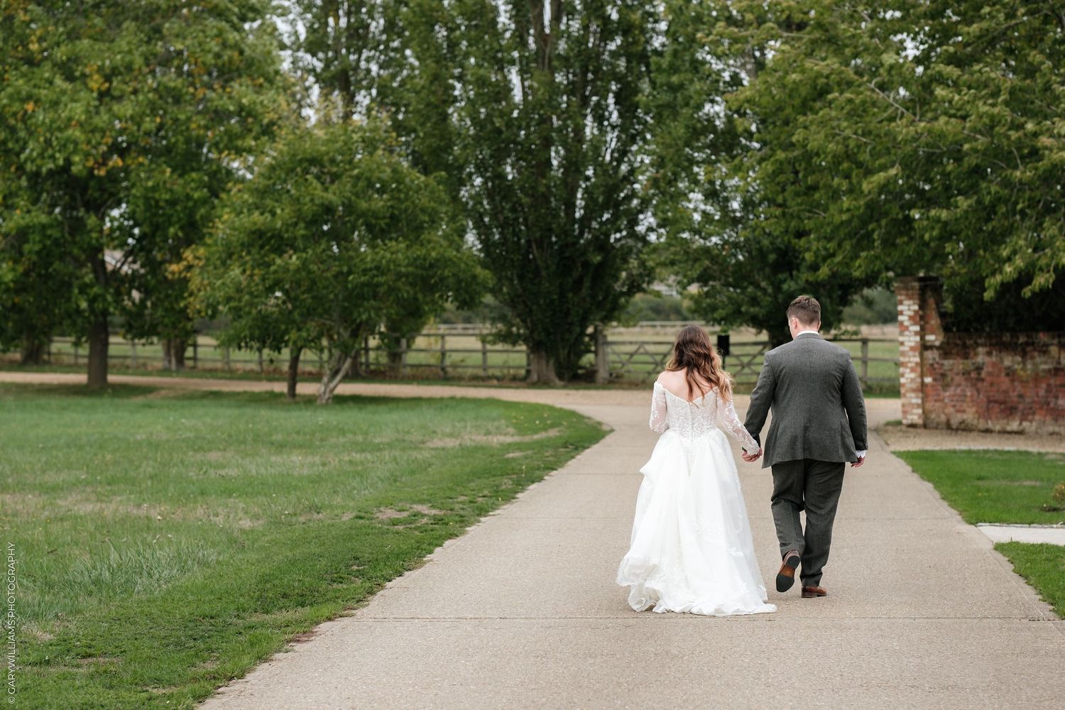 A bride and groom walk hand in hand down a curved path surrounded by lush green trees on their wedding day.