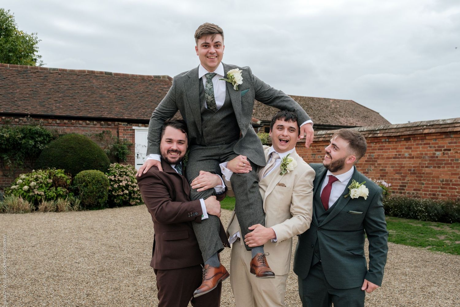 Groomsmen lifting groom on shoulders outdoors at rustic wedding venue, all wearing suits with boutonnieres.
