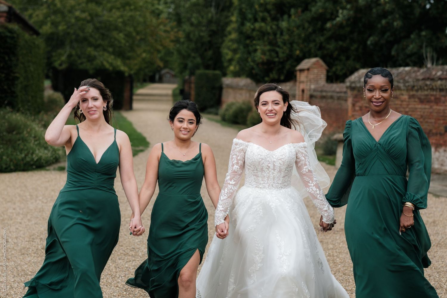 Bride in white lace gown walks with three bridesmaids in emerald green dresses along a gravel path.