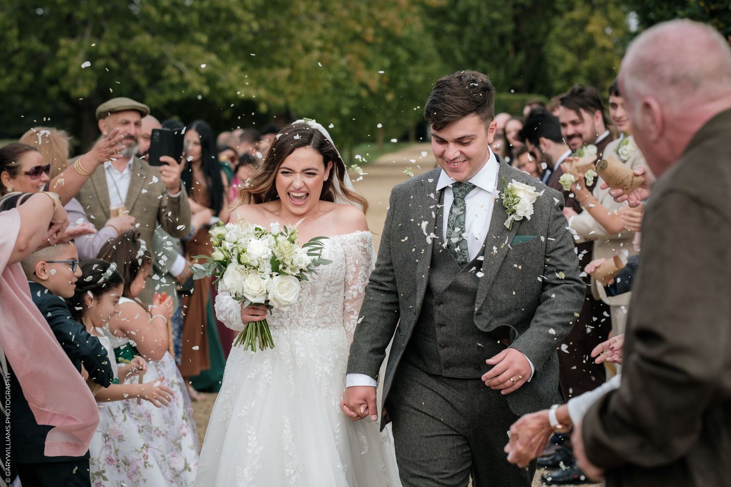 Bride and groom walk through confetti shower surrounded by cheering guests at outdoor wedding ceremony.