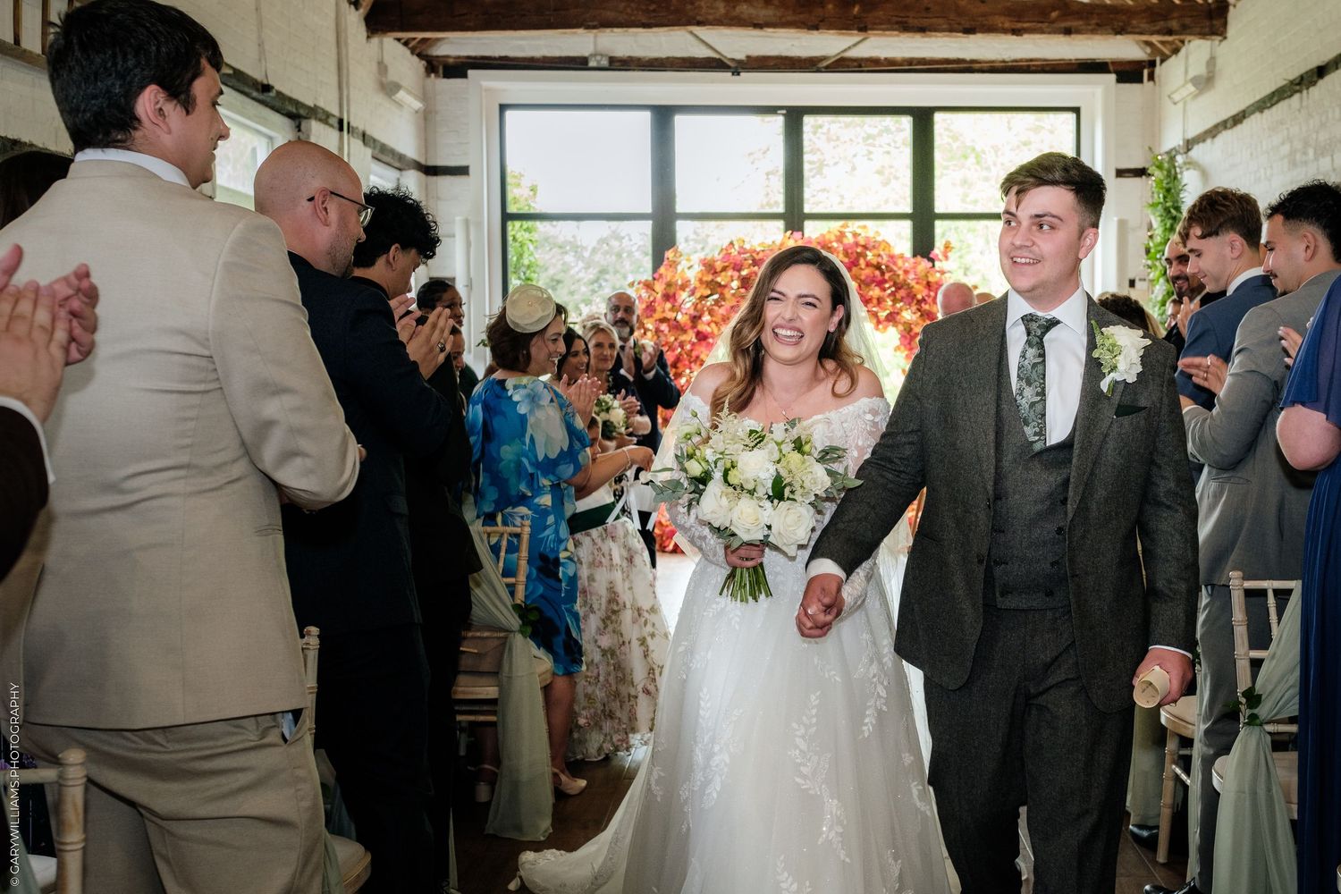 Bride and groom walk down the aisle smiling, guests applauding in a decorated indoor wedding venue.