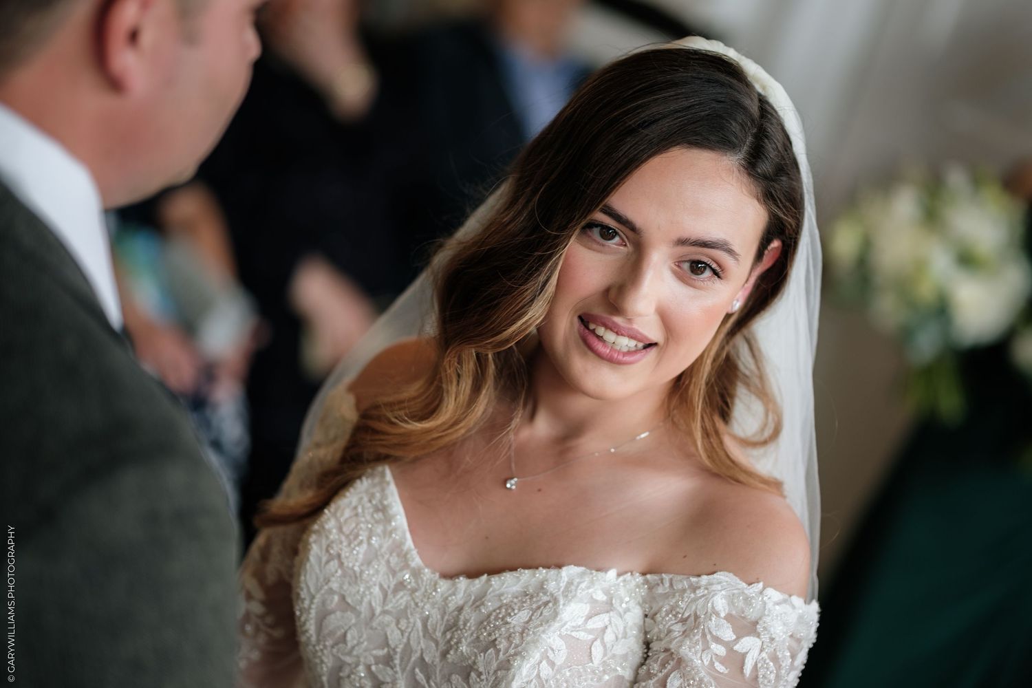 Bride in lace off-shoulder gown and veil smiles warmly during wedding ceremony.