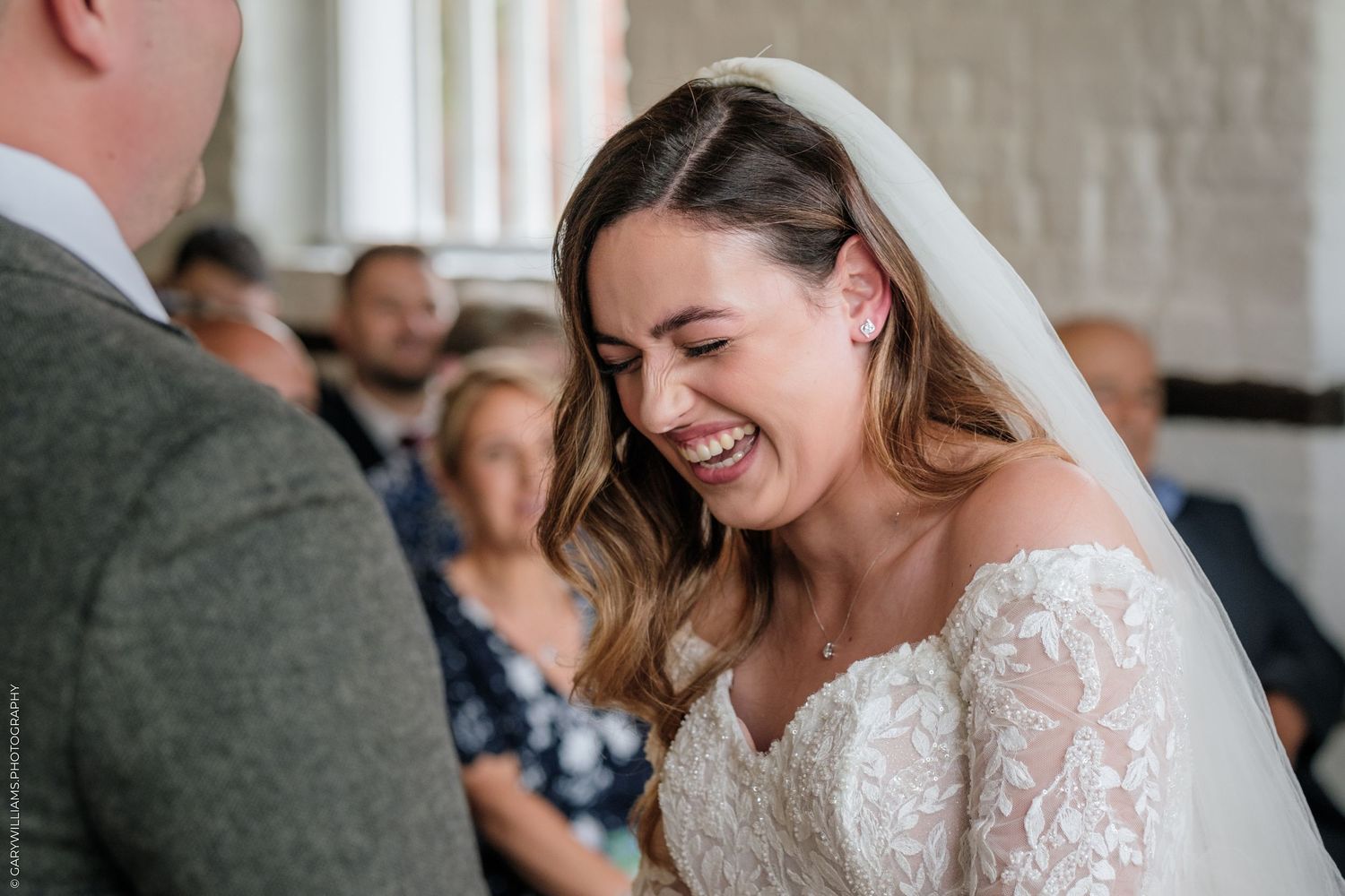 A joyful bride in a lace gown and veil laughs during her wedding ceremony indoors.