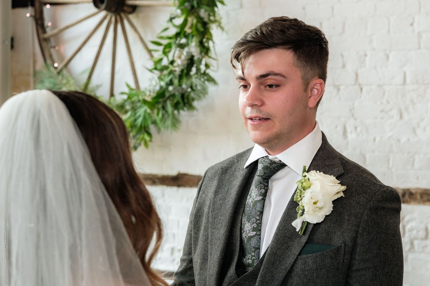 Groom in gray suit with white boutonniere smiling at bride in veil during wedding ceremony.