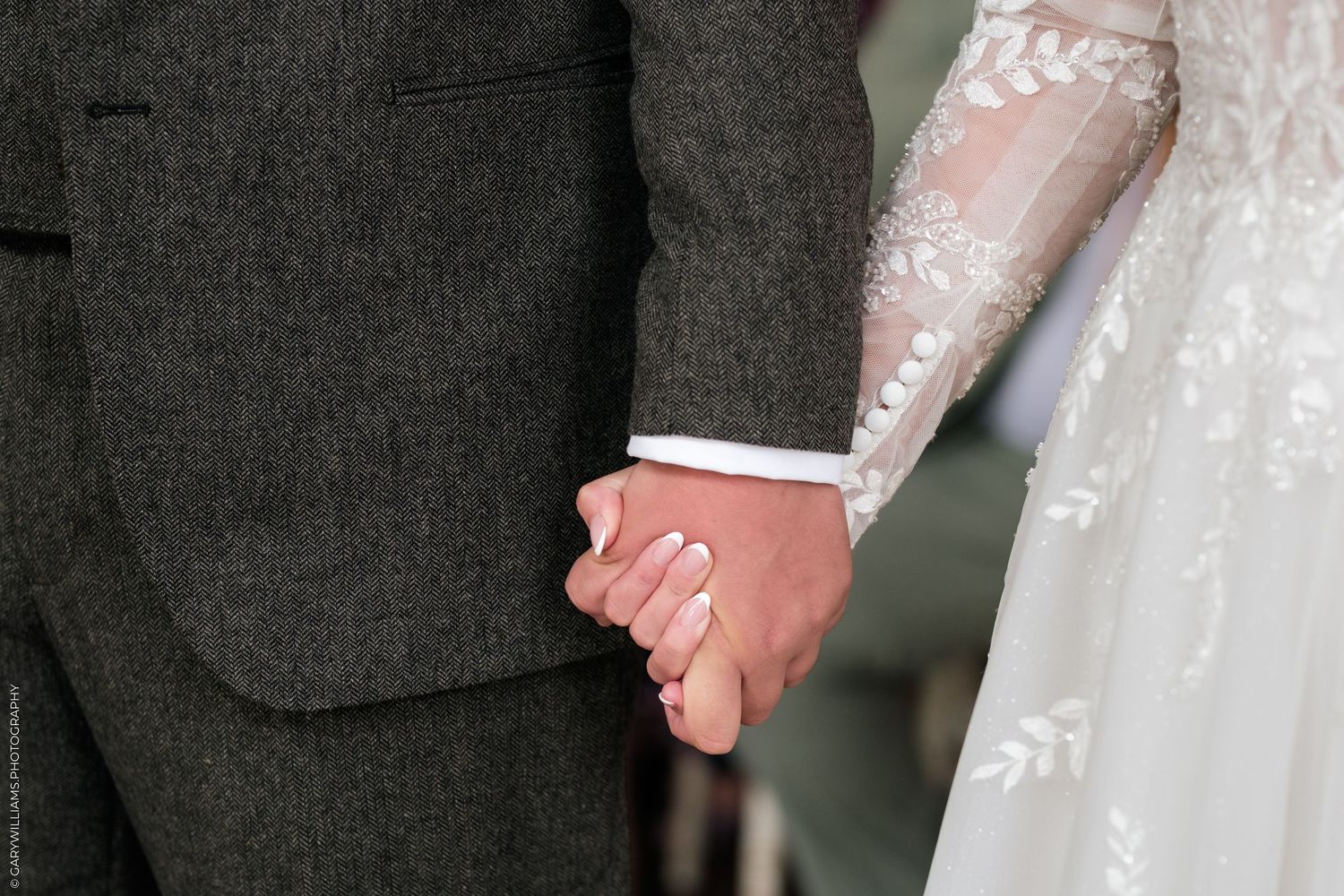 Close-up of a bride and groom holding hands, showcasing lace wedding dress sleeve and gray tweed suit.