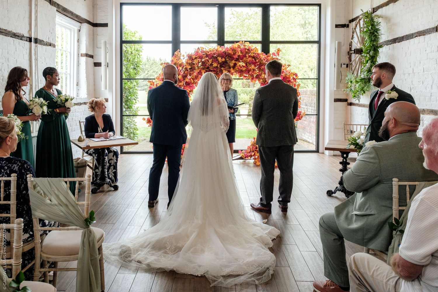 Bride and groom stand at floral arch altar during elegant indoor wedding ceremony with guests seated in white chairs.