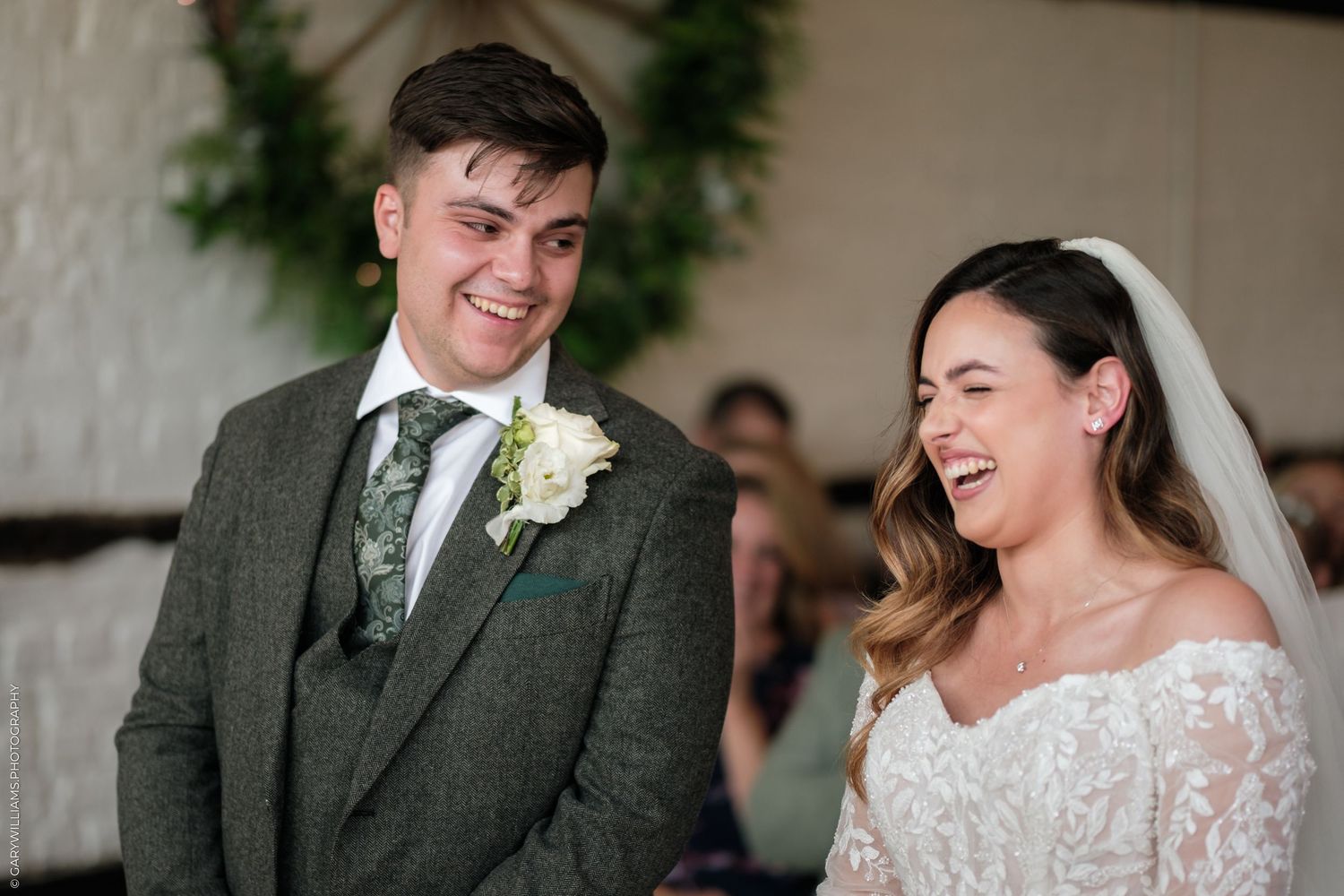 Bride and groom laughing joyfully during wedding ceremony, groom in green tweed suit, bride in lace dress with veil.