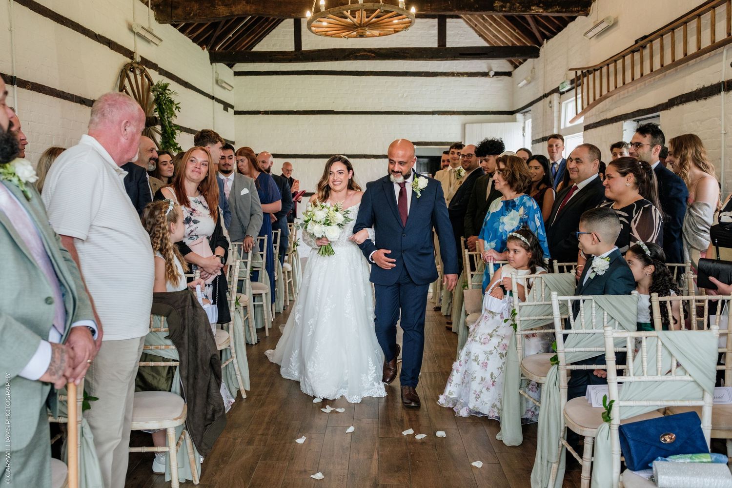 Bride and groom walk down the aisle at an indoor wedding ceremony, surrounded by seated guests in a bright venue.