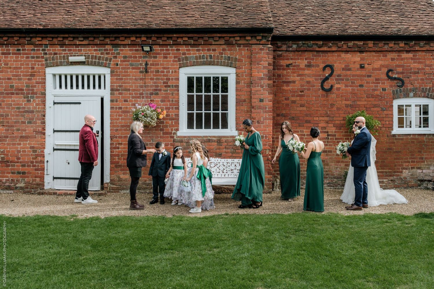 Wedding party in green dresses poses outside a rustic red brick venue with bride and groom on a sunny day.