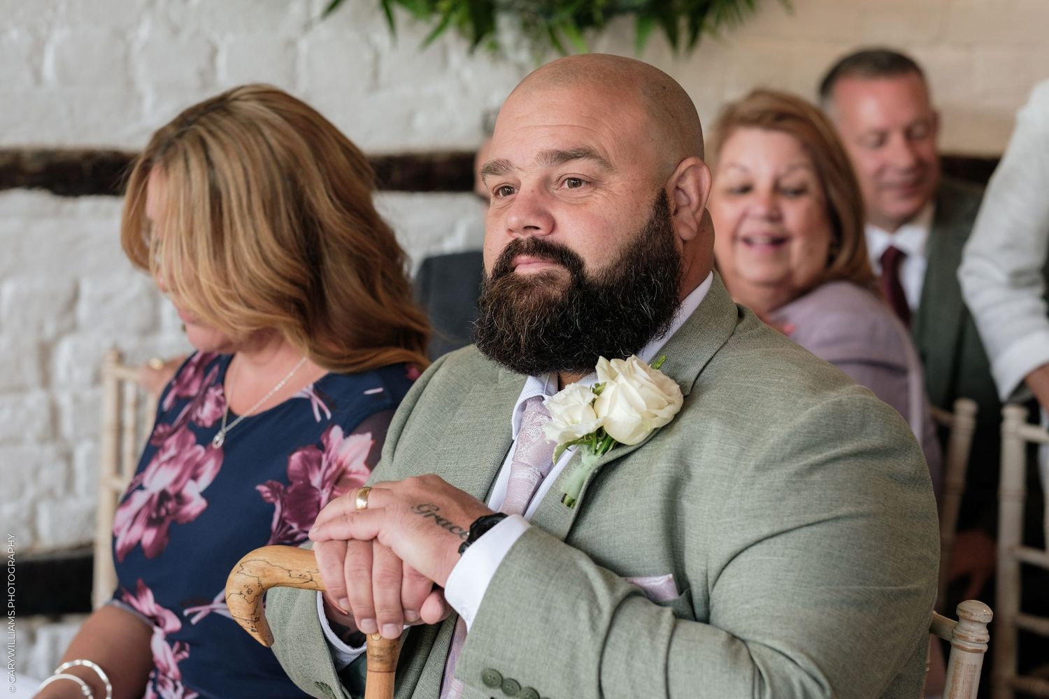 Bald bearded man in sage green suit with white boutonniere sits at wedding reception holding a wooden cane.