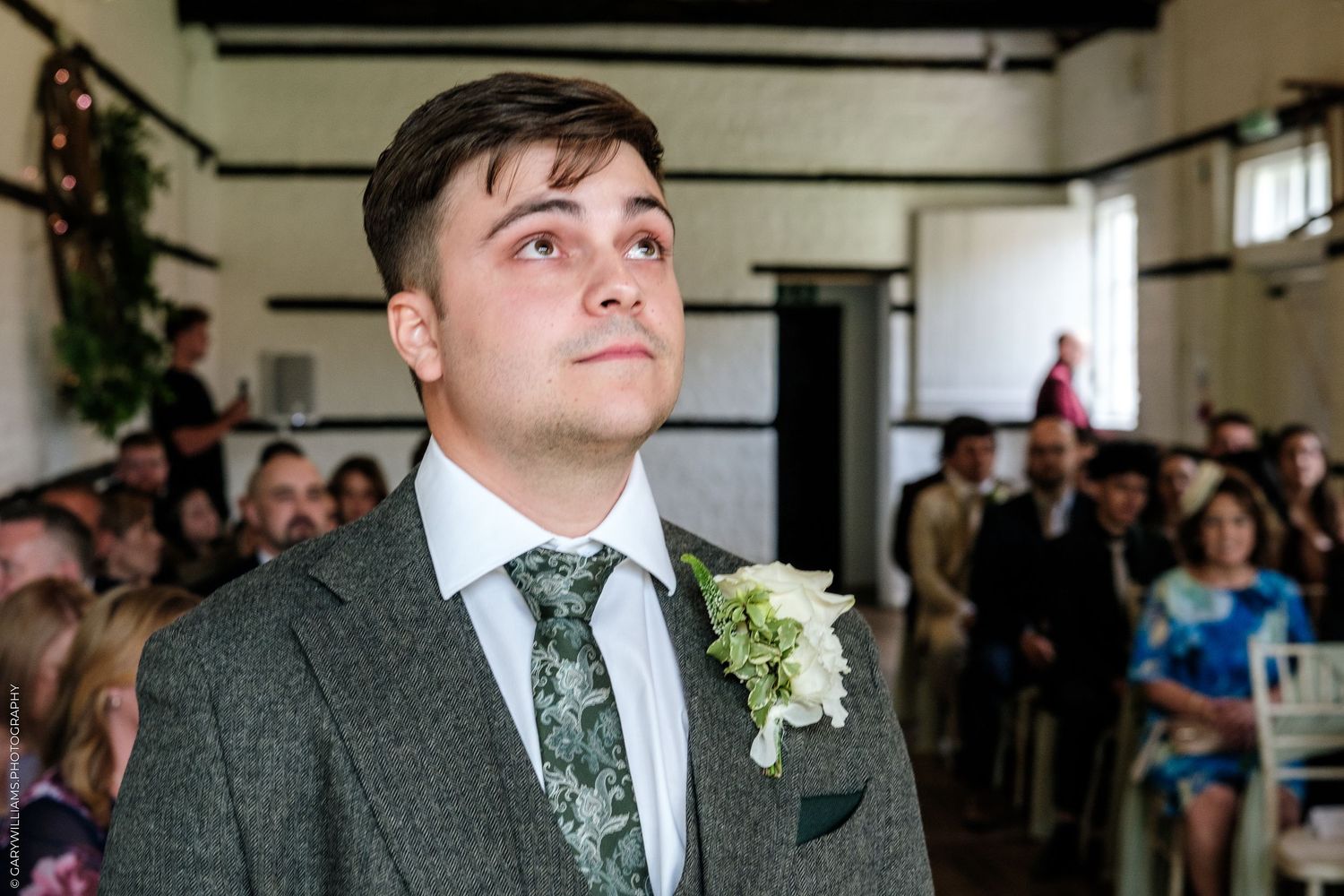 Young groom in grey suit with floral boutonniere gazes upward emotionally during wedding ceremony.