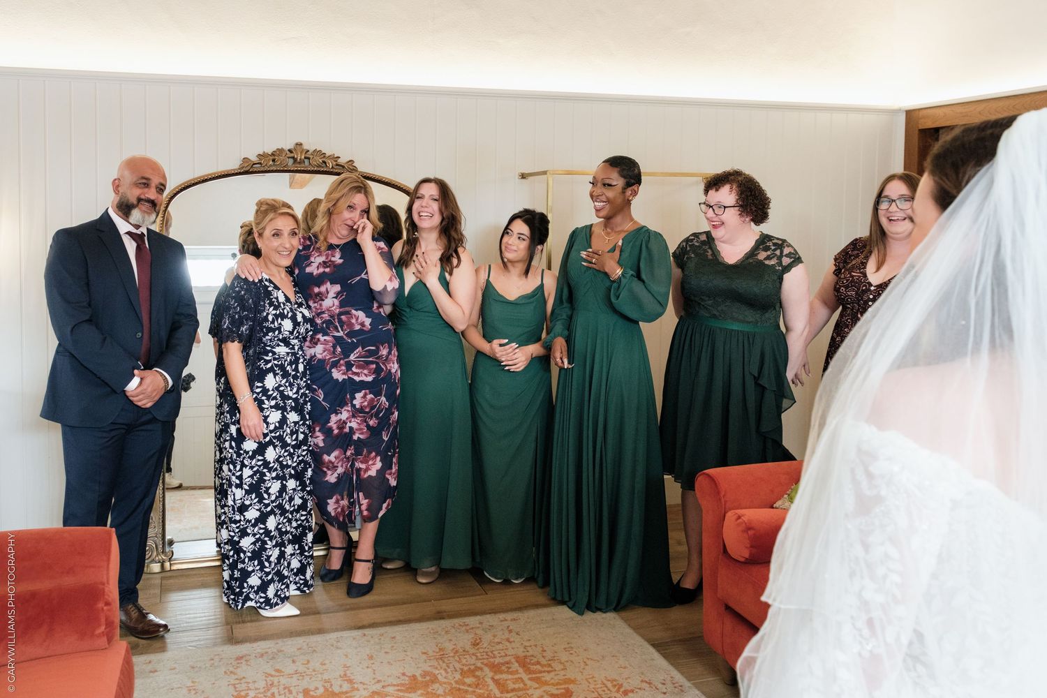 Bridal party in green dresses reacting emotionally as bride reveals wedding gown in elegant room.