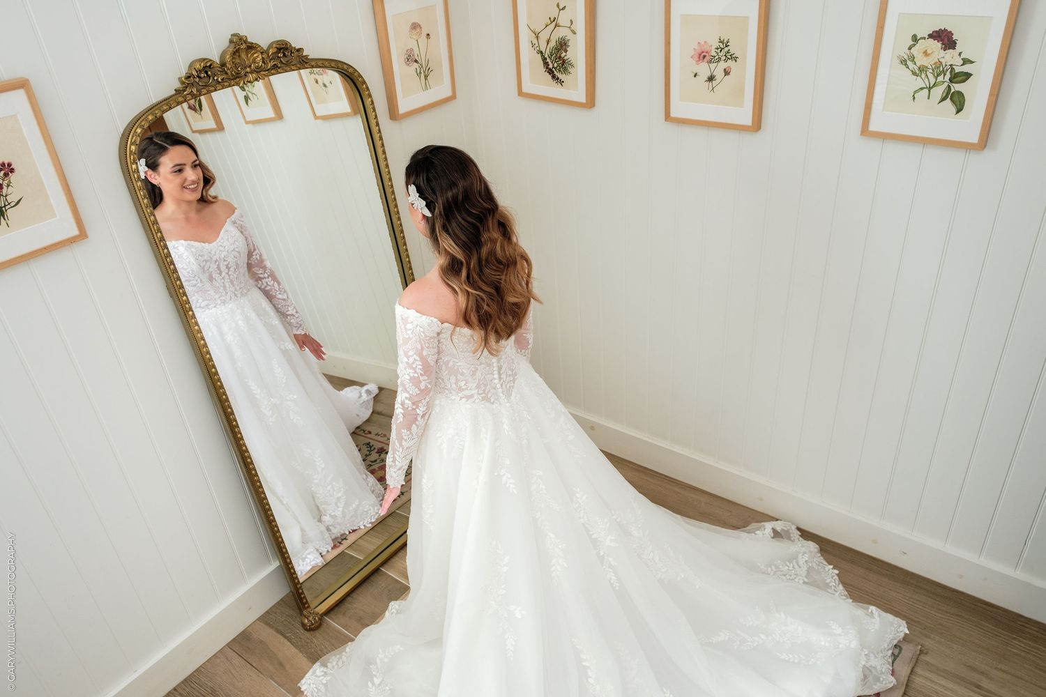 Bride in elegant lace wedding gown admiring her reflection in an ornate gold floor mirror in a bright white room.