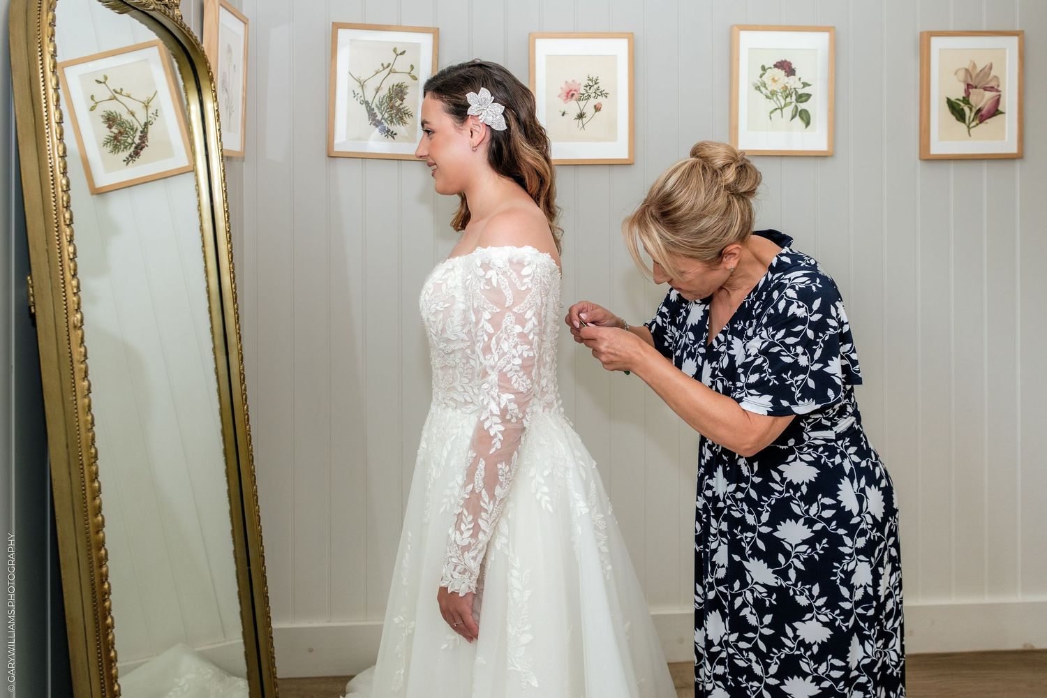 A woman in a lace wedding gown gets her dress fastened by another woman in a floral dress near a gold mirror.