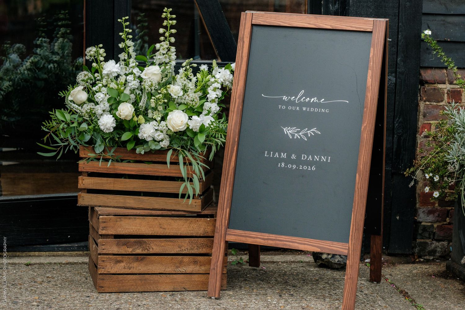 Elegant wedding welcome chalkboard sign for Liam and Danni beside white floral arrangement in wooden crate.