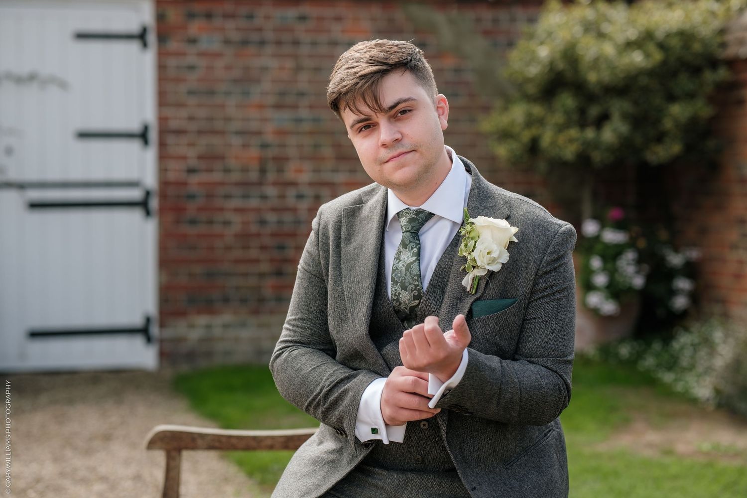 Young groom in grey tweed suit holding boutonniere while seated outdoors near rustic brick building.