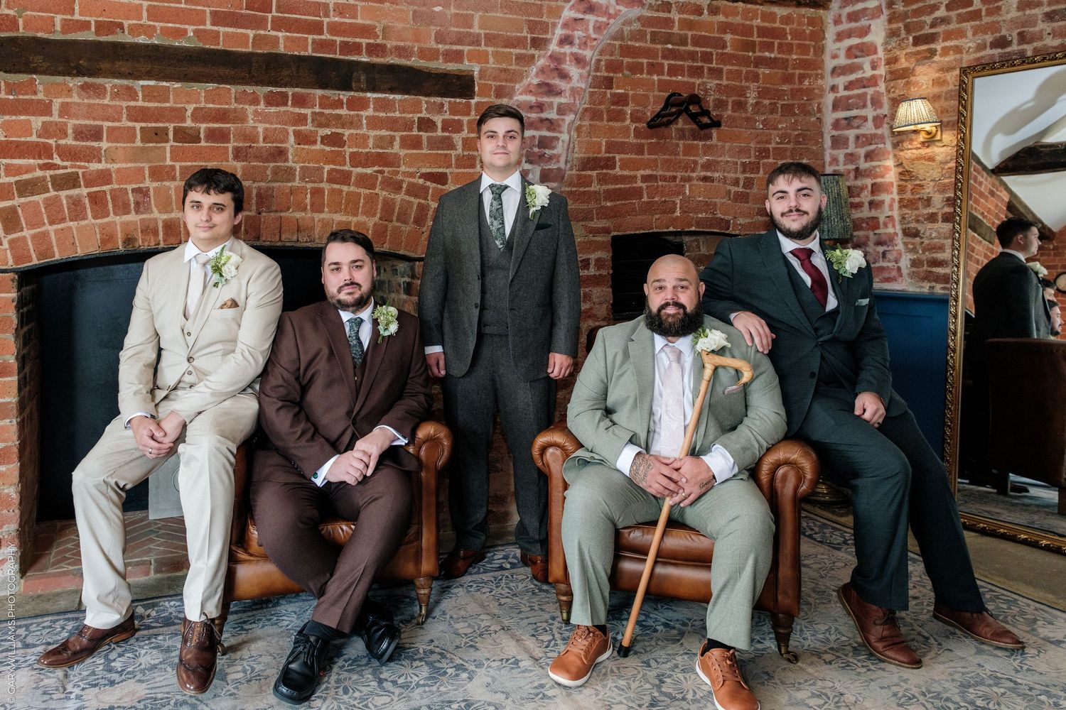 Five groomsmen in suits pose in a rustic brick venue, mixing tan, brown, and green attire for a stylish wedding party photo.
