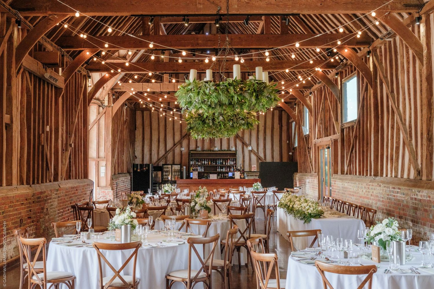 Rustic barn wedding reception with string lights, greenery chandelier, white-draped round tables, and wooden cross-back chairs.