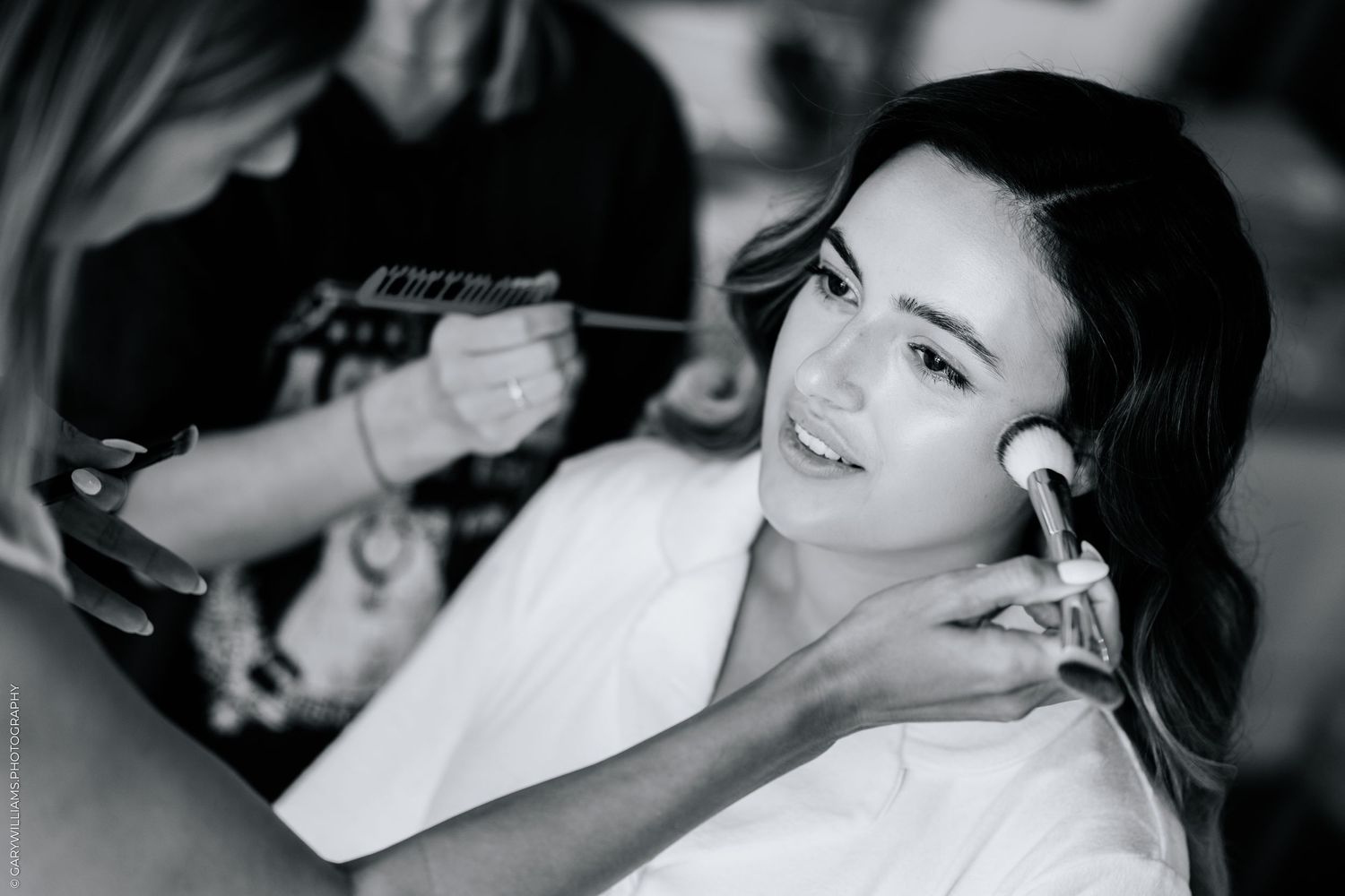 Black and white photo of a bride getting makeup applied by two artists before her wedding ceremony.