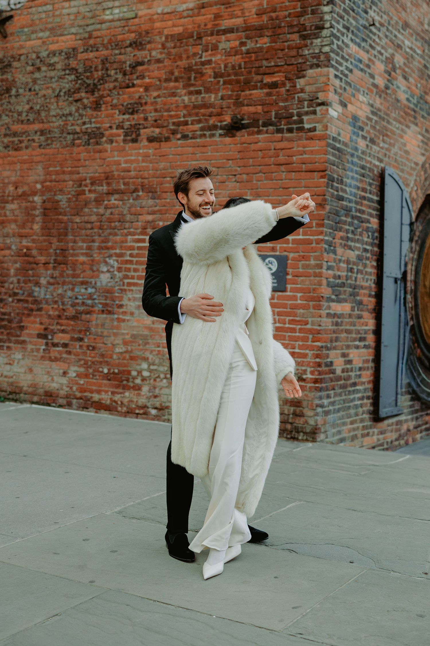 A joyful couple dances outdoors, the bride in a white fur coat and pantsuit, groom in black suit, against a brick wall.