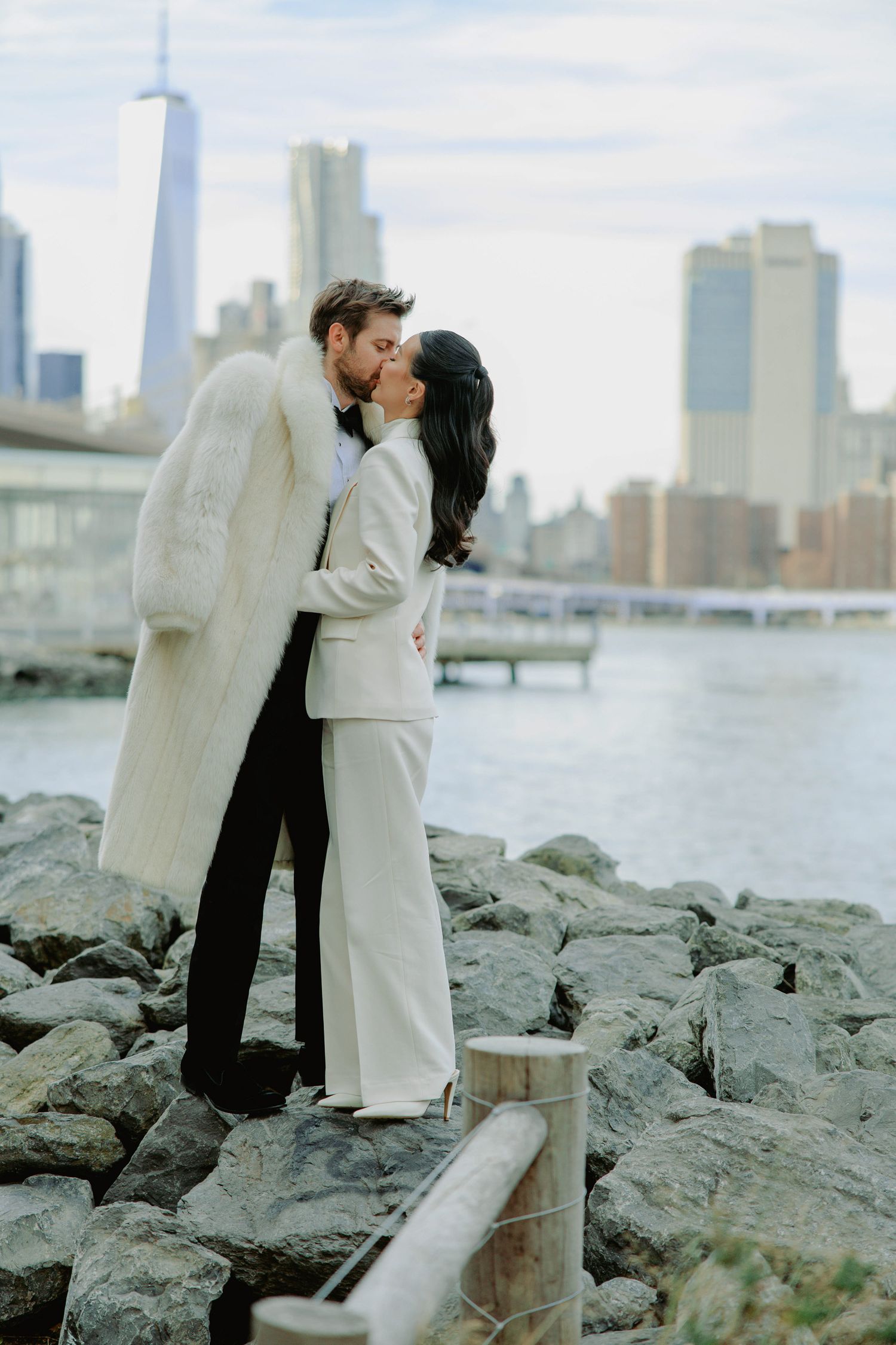 Couple kissing in white outfits on rocky NYC waterfront with Manhattan skyline and One World Trade Center in background.