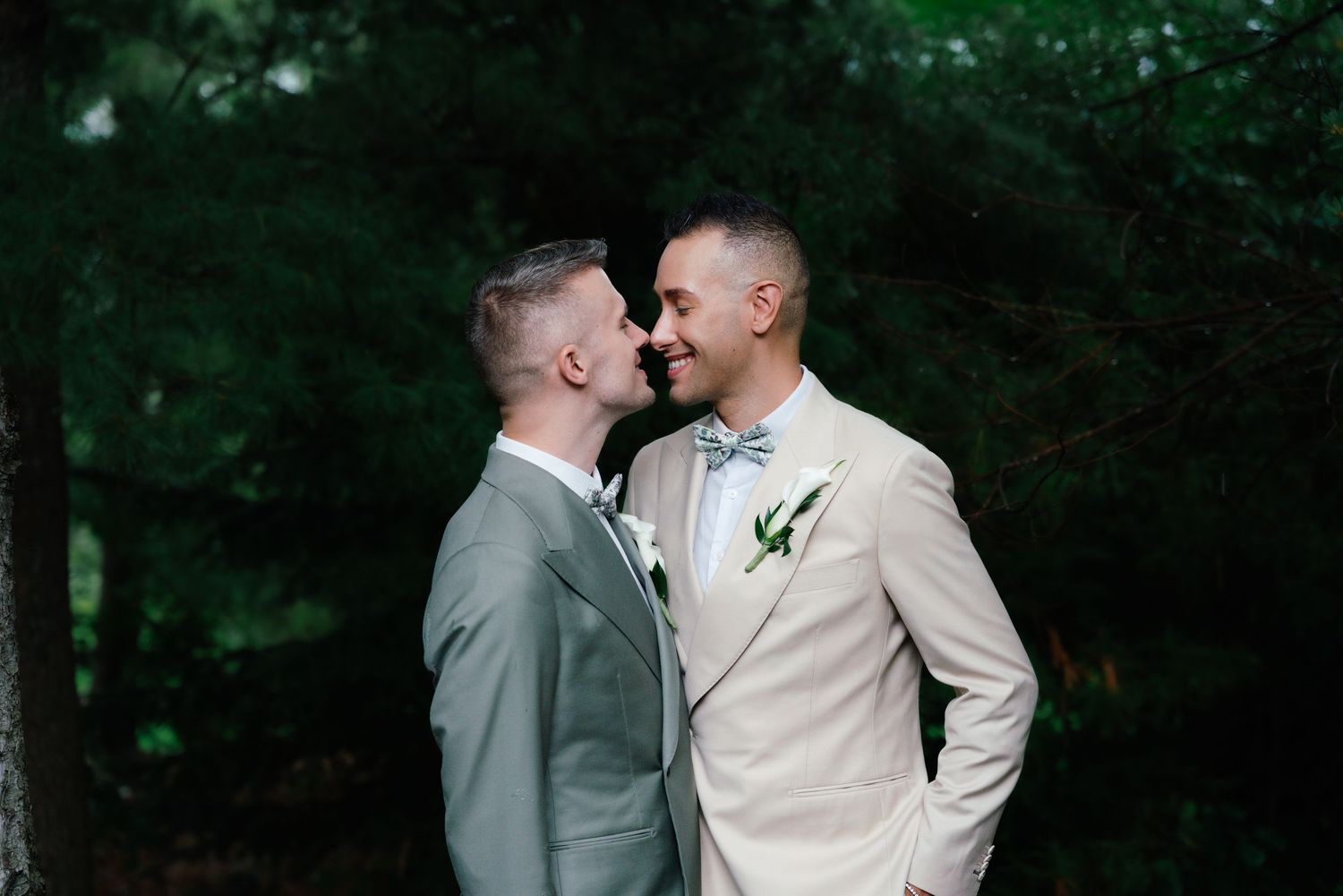 Two grooms in suits share a romantic moment outdoors, surrounded by lush green trees on their wedding day.