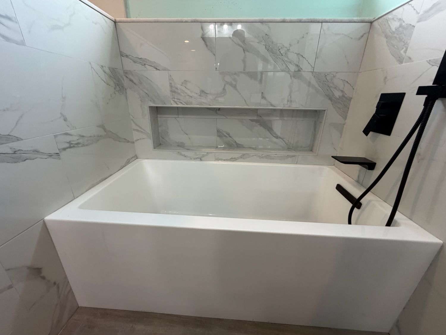 Modern white freestanding bathtub with marble tile surround and built-in niche, featuring matte black fixtures.