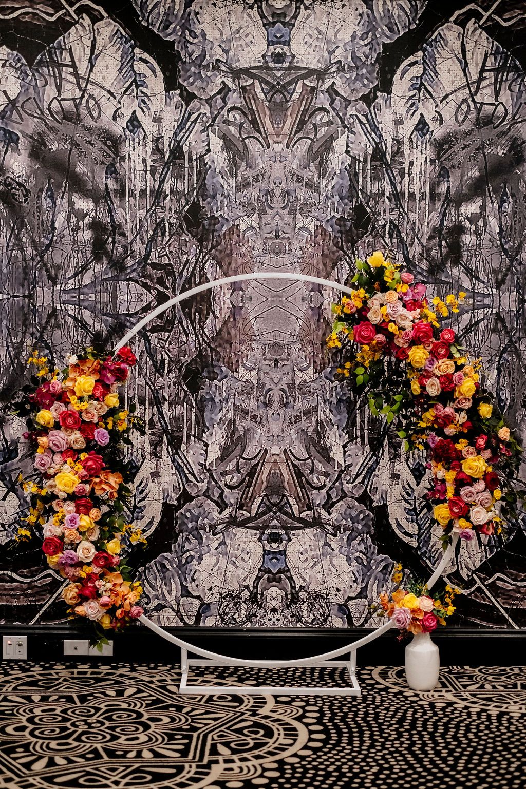 Circular white arch adorned with vibrant yellow, pink, and red floral arrangements against an ornate black and white wallpaper backdrop.