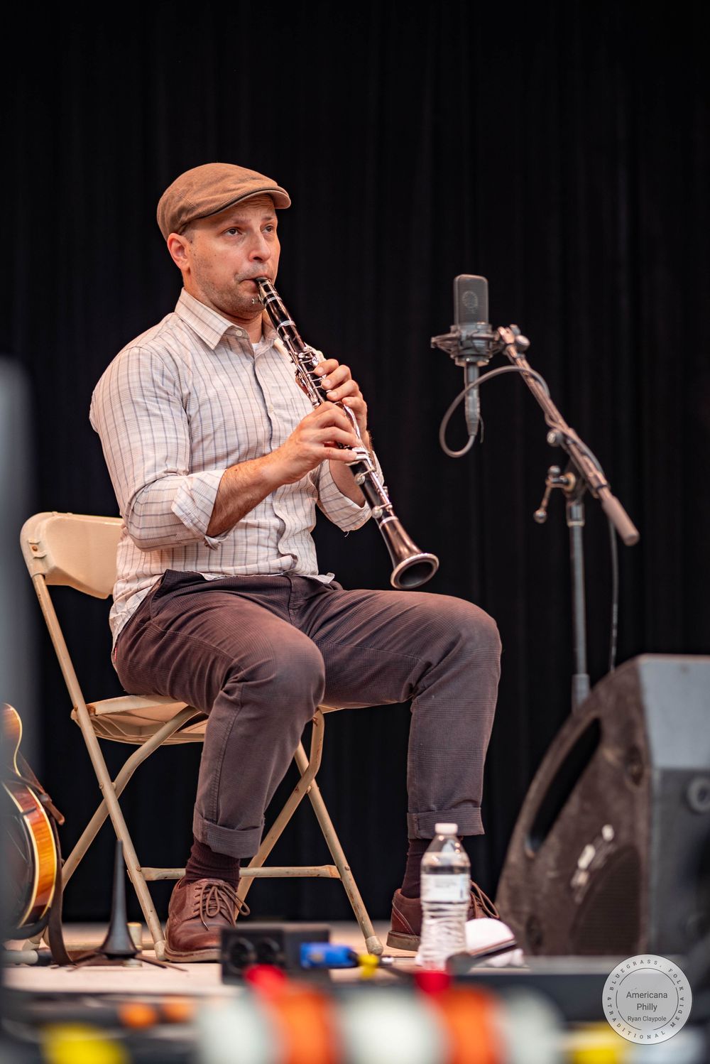 Musician playing clarinet seated on folding chair on stage with microphone stand nearby.