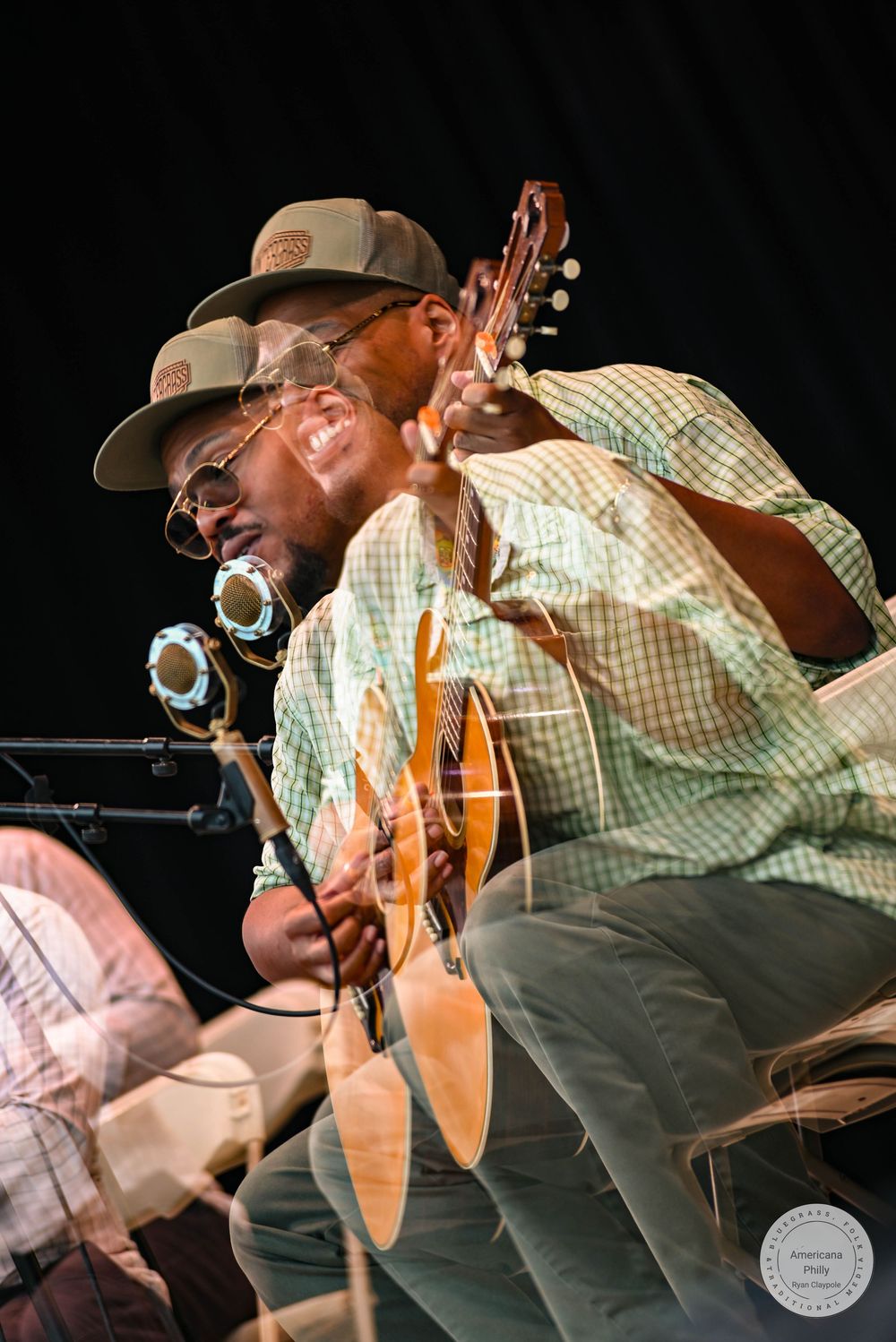 A musician smiles while playing acoustic guitar on stage, wearing a cap and headphones around his neck.